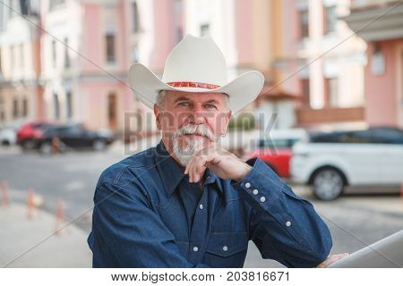 A mature cowboy in a hat jeans and a denim shirt looks at the camera. On open air
