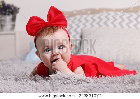 Funny baby in red dress and bow with hand in mouth is on bed with pillows in bedroom