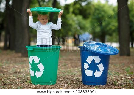 Blue and green bins. Cute preschooler boy, looking inside the trash bin in the street, summer outdoor, save the planet, save the Earth concept. Day in park. Funny babies.