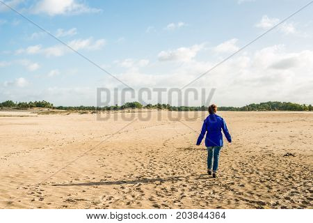 Woman in a blue jacket walks into a nature area with lots of sand. It's a sunny day in summer.