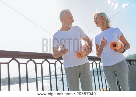 Enjoy training together. Cheerful athletic elderly couple walking down the bridge and holding yoga mats while planning to practice yoga
