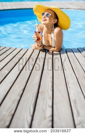 Young woman in swimsuit with big yellow sunhat relaxing with a bottle of fresh drink sitting on the poolside outdoors