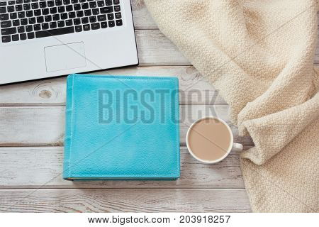 Female workplace with a laptop cup of coffee blue notebook and cozy sweater on light wooden background.Top view composition with copy space.