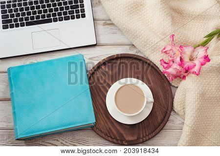 Female workplace with a laptop cup of coffee blue notebook cozy sweater and pink flowers on light wooden background.Top view composition with copy space.