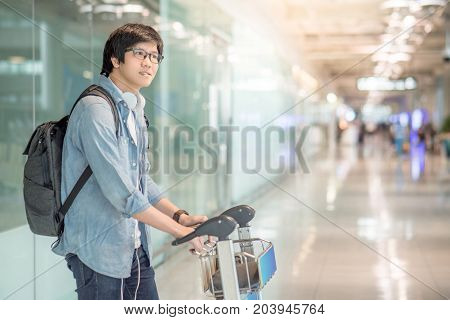 Young Asian man walking with airport trolley and his suitcase luggage in the international airport terminal arrival from travel abroad