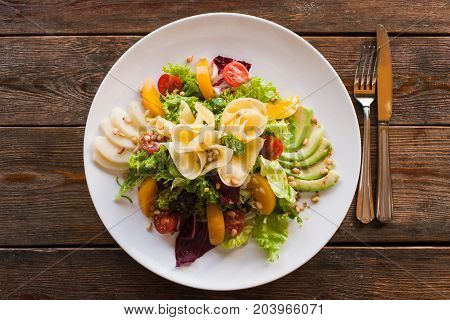 Lettuce salad with cheese and fruits on plate. Fresh vegetables decorated pine nuts, avocado, pearch and pear, serving in restaurant, top view