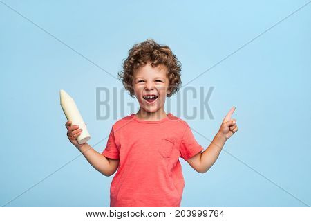 Adorable curly kid in casual clothing holding mockup big pencil looking at camera on blue background.