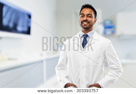 medicine, dentistry and profession concept - smiling indian male dentist in white coat over dental clinic office background