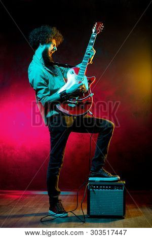 Portrait Of Hipster Man With Curly Hair With Red Guitar In Neon Lights. Rock Musician Is Playing Ele