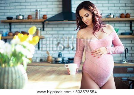 Young Pregnant Woman In Pink Bodysuit Drinking Water In The Kitchen. How To Get Rid Of Morning Sickn