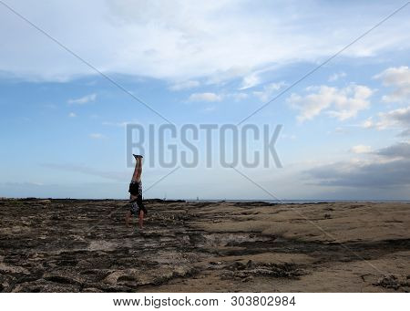 Man Wearing A T-shirt, Shorts, And Slippers Handstands On Shore Rocks Of Ko Olina On Oahu, Hawaii Wi