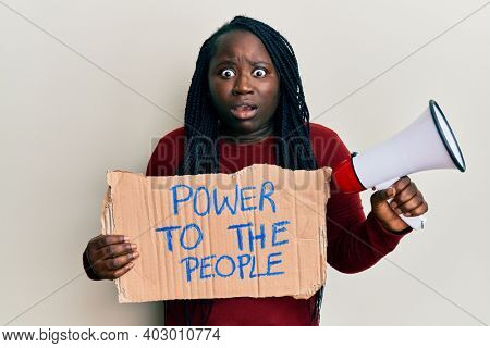Young black woman with braids holding power to the people banner and megaphone afraid and shocked with surprise and amazed expression, fear and excited face. 