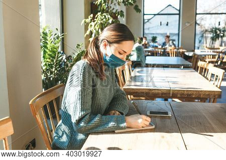A Girl In A Protective Mask Writes In A Notebook With A Pen. She Is Learning Or Writing Goals Or Som