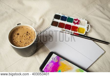 Set Of Watercolor Paints, Scetchbook, Paintbrush And A Cup Of Coffee For Breakfast.
