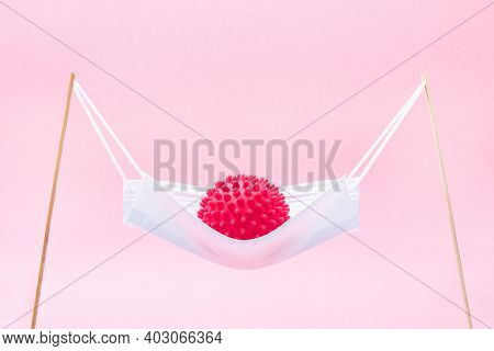 A Red Ball With Spikes Coronavirus Swinging In A Medical Mask Like In A Hammock Or Swing.