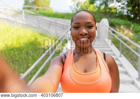 fitness, sport and technology concept - happy smiling young african american woman with wireless earphones listening to music and taking selfie outdoors