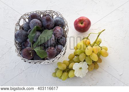 Just Picked Ripe Plums In A Wicker Basket, An Apple And A Bunch Of Ripe White Grapes On The White St