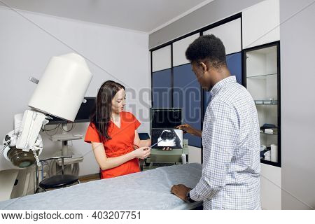 Young Female Caucasian Doctor In Modern Clinic, Holding Tablet Pc And Showing To Her Male African Pa