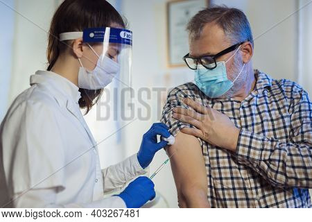 Young Woman Nurse With Surgical Mask And Face Shild Giving Injection To Senior Man. Covid-19 Or Coro