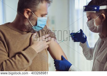 Young Woman Nurse With Surgical Mask And Face Shild Giving Injection To Senior Man. Covid-19 Or Coro