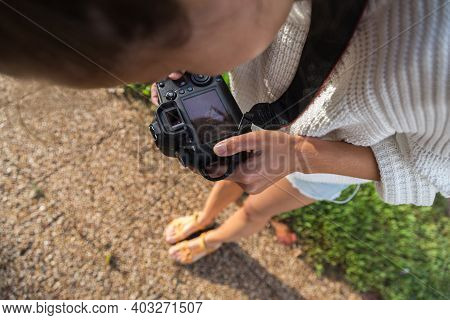 A Girl With Reflex Camera Is Taking Photos Of Her Legs In Sandals, Standing On Sidewalk With Green L