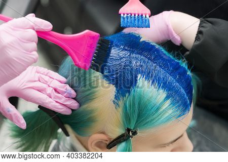 Two Hairdressers Using Pink Brush While Applying Blue Paint To Female Client With Emerald Hair Color