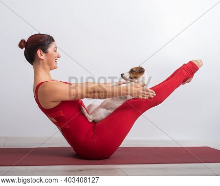 A Woman In A Red Overalls Practices Yoga With A Dog. The Girl Performs The Vi Balance Asana On A Whi
