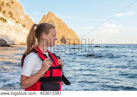Teenage Girl In Red Life Jacket Stands On Ocean And Looks At Sea. Means Of Rescuing People On Water,