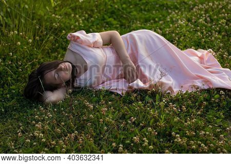 A Girl With Dark Hair In A Long Pink Dress Lies And Sleeps In The Green Grass With White Clover Flow