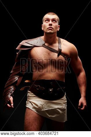 Gladiator With Sword And Armor On A Black Background. A Warrior In Gladiatorial Armor Looks Up At A 