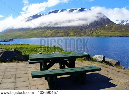 An Empty Picnic Table Overlooking The Snowy Mountain And Blue Fjord In The Summer Near Olafsfjordur,