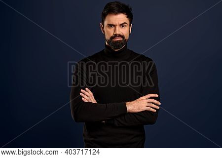 Portrait Of Serious Displeased Male Stands Crossed Hands Against Blue Background, Scowl Face In Diss