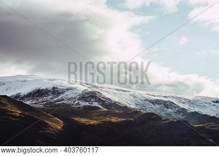 Atmospheric View To Snowy Mountains In Sunlight Under Cloudy Sky. Low Clouds Among Big Rocks With Sn