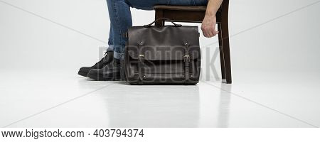 Man In A Blue Jeans And Black Boots Sits On A Chair With A Brown Mens Shoulder Leather Bag For A Doc
