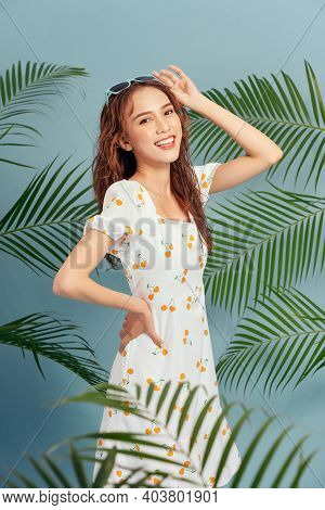 Portrait Of Stylish Serious Girl Wears Summer Dress Bahind The Tropical Leaves Over Blue Background.
