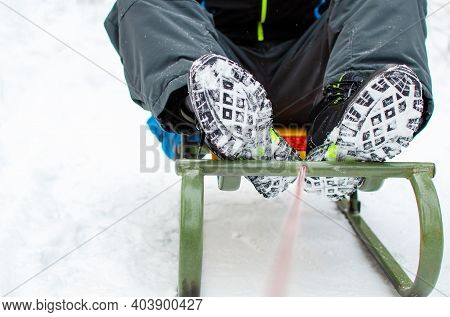 Winter Holidays And Games Concept. The Child Is Sitting On A Sleigh. Close-up Legs In The Snow. Wint