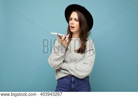 Young Asking Dissatisfied Brunette Woman Speaking Wearing Black Hat And Grey Sweater Holding Smartph