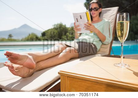 Woman reading book by swimming pool with champagne on table in foreground