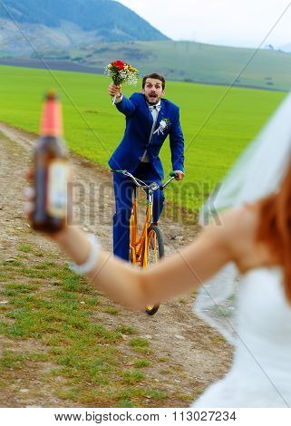 drunken groom on a bike holding a wedding bouquet is running after a bride with a beer bottle.