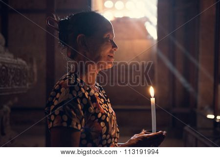 Old wrinkled traditional Asian burmese woman praying with candle light inside a temple, low light, Myanmar
