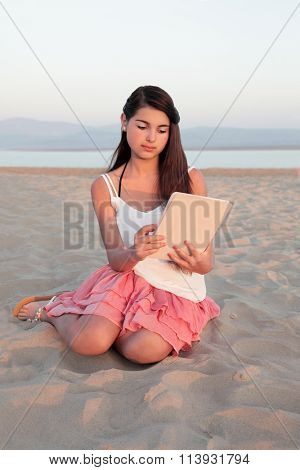 Young european teenage girl with long dark hair browsing on a computer tablet device on a beach in summer.