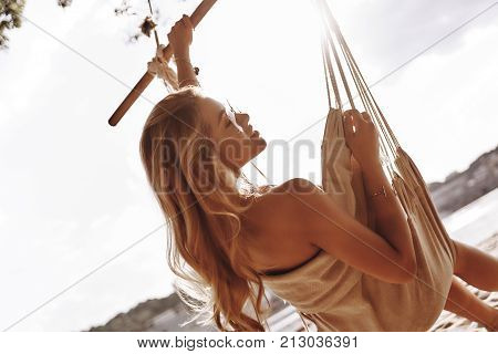 Enjoying warm day. Rear view of attractive young woman in swimwear sunbathing while sitting in the hammock on the beach