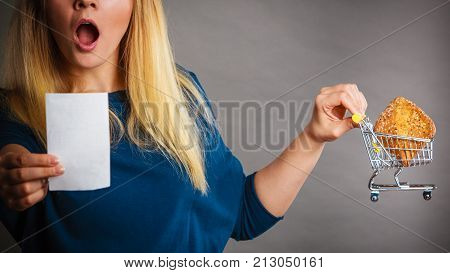 Shocked woman holding shopping basket with bread looking at bill receipt being scared of huge prices