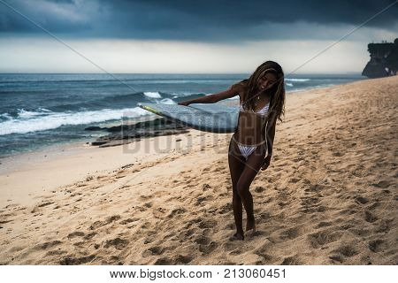 Young smiling dark skin woman in bikini carrying surfboard at beach in rainy day
