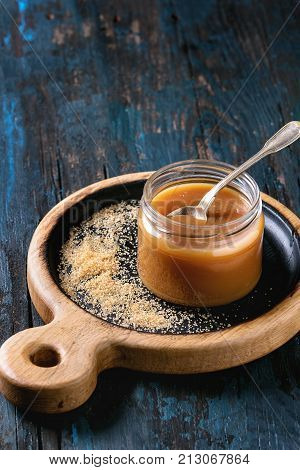 Glass jar of homemade salted caramel sauce on wooden serving board with ingredients brown sugar above. Over old dark blue wooden background.