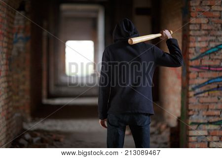 A street hooligan in a black hood on a construction site stands with a wooden bat on his shoulder. Red brick tunnel.