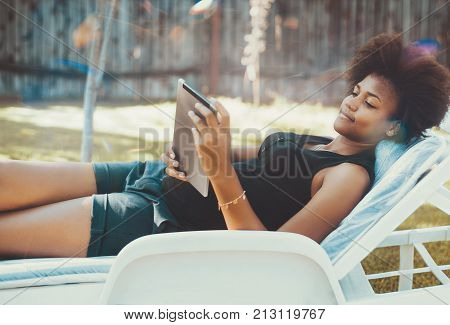 Thoughtful black teenage female on daybed in public park is surfing the net with digital pad; young beautiful African American girl is chilling on deck chair with tablet pc in the summer garden