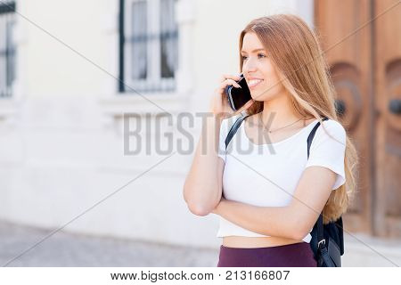 Smiling young woman communicating on phone and looking with confidence straight. Cheerful successful girl with red hair talking on cell phone while walking over city. Modern life concept