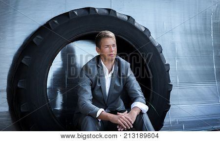 Close up outdoor portrait of confident serious young Caucasian student wearing grey stylish formal suit relaxing looking afar. Handsome successful male sitting inside tyre taking rest after walking.