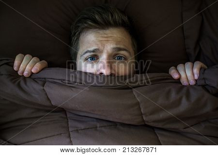 Caucasian young man hiding in bed under the blanket at home. He is stressed and tries run away from problems